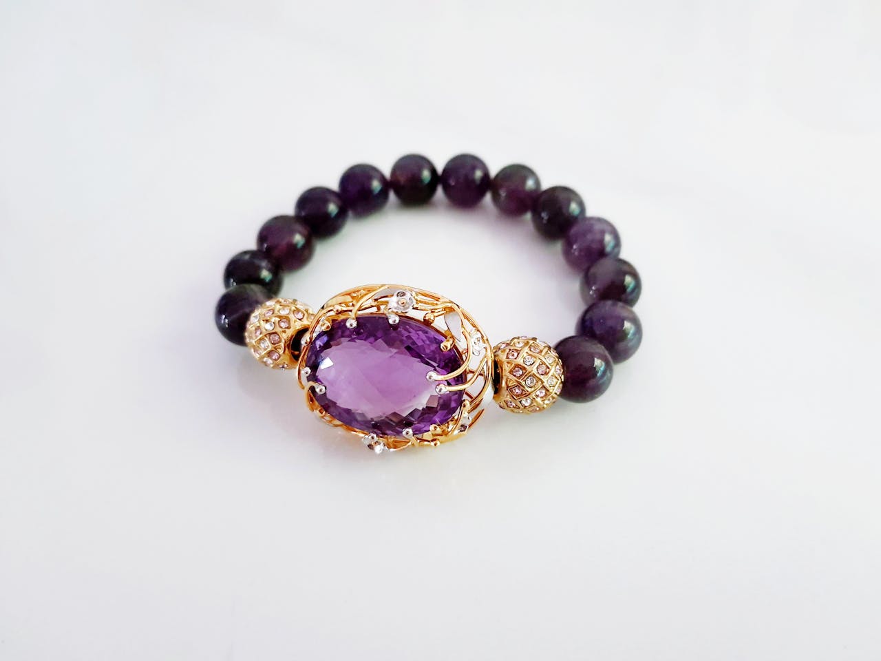 about-us-02 Luxurious beaded bracelet with central amethyst gemstone set in gold, on a white background.